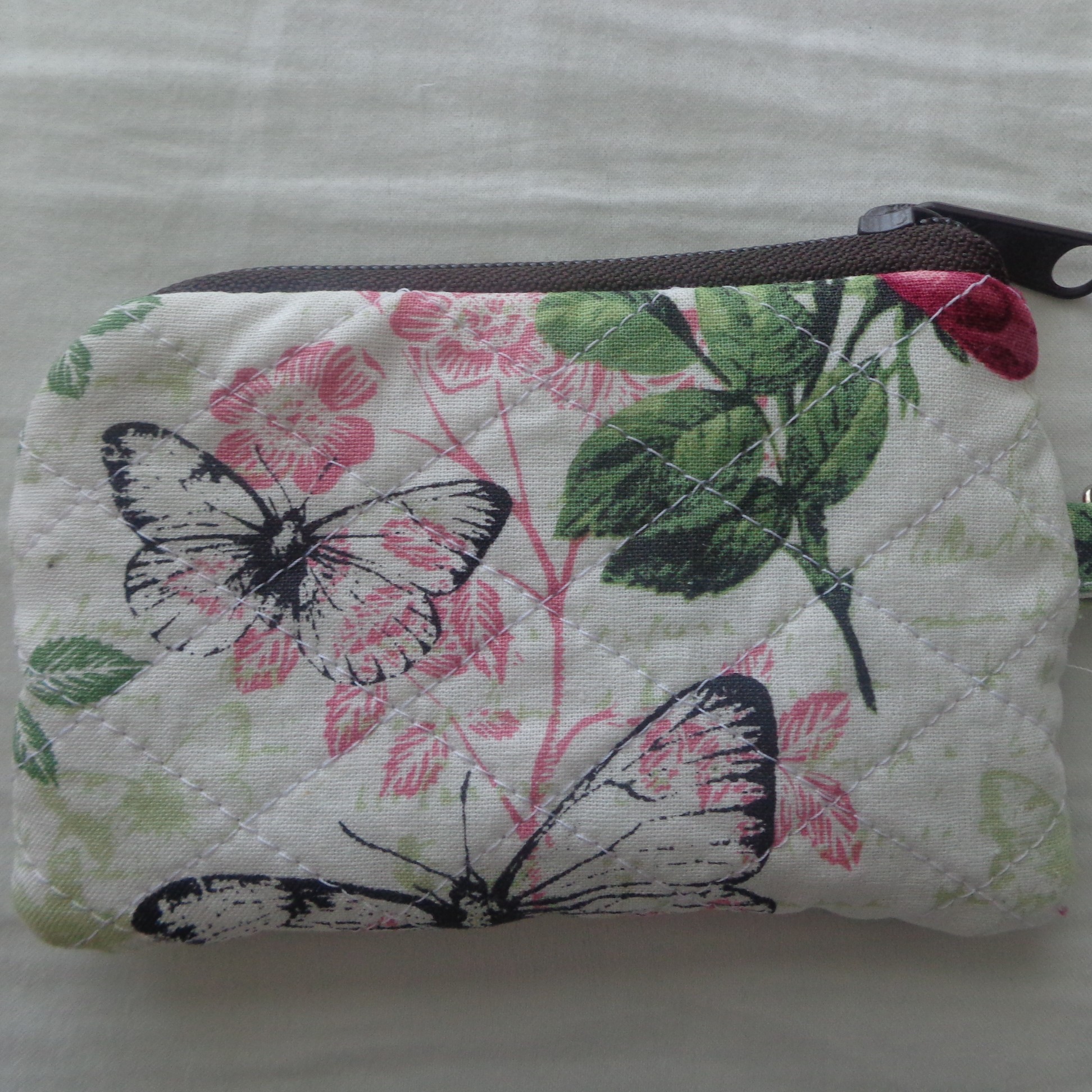 a fist sized handsewn pouch with a brown zipper and a cream fabric that has black butterflies, pink flowers, and green leaves printed across it.